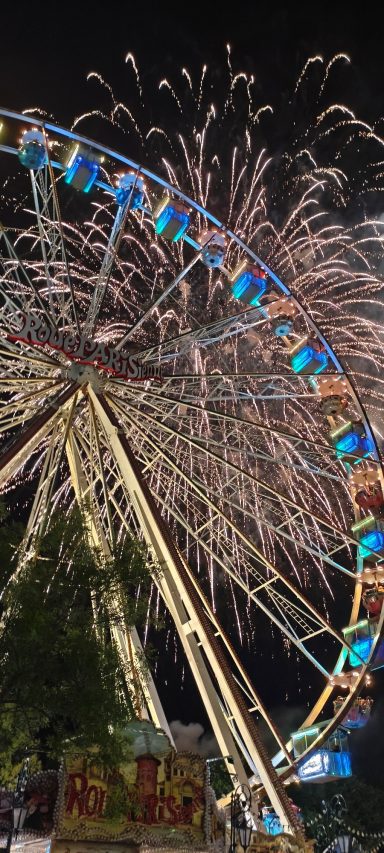 Riesenrad bei Nacht, umgeben von bunten Feuerwerken.