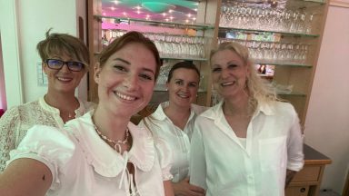 Vier Frauen in hellen Outfits lächeln in die Kamera, stehen nebeneinander.