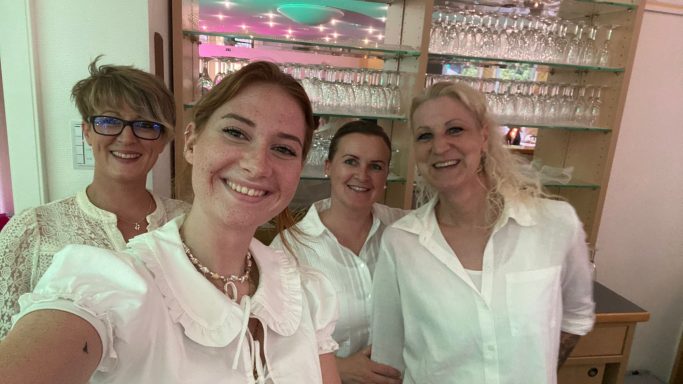 Vier Frauen in hellen Outfits lächeln in die Kamera, stehen nebeneinander.