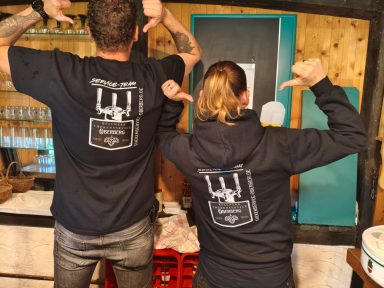 Zwei Personen mit schwarzen T-Shirts, die nach hinten zeigen, in einem rustikalen Ambiente.