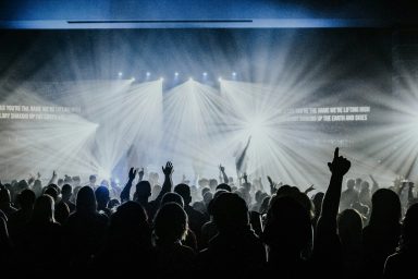 Menschenmenge mit erhobenen Händen auf einem Konzert, umgeben von Lichtstrahlen.