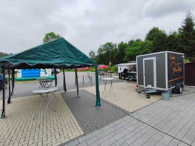 Blick auf einen Stand mit Zelt, Tischen und einem Verkaufswagen auf gepflastertem Boden.
