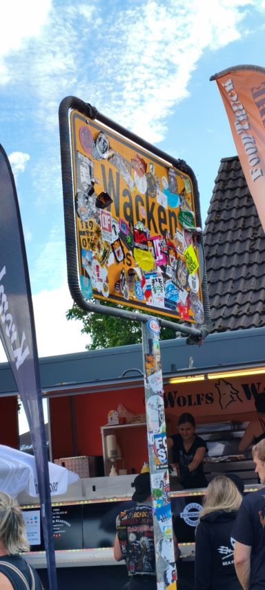 Bunte Verkehrsschilder mit Stickerkunst vor einem Verkaufsstand und Menschenansammlung.
