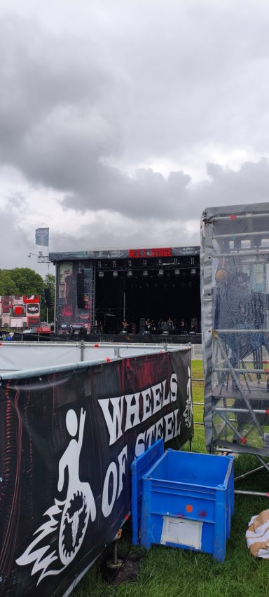 Bühne mit Blackout-Vorhang und blauen Kisten auf einem Festivalgelände bei bewölktem Himmel.