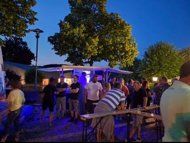Personen stehen an Tischen bei Abendveranstaltung unter Bäumen mit blauer Beleuchtung.