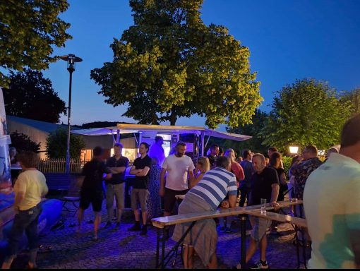 Personen stehen an Tischen bei Abendveranstaltung unter Bäumen mit blauer Beleuchtung.