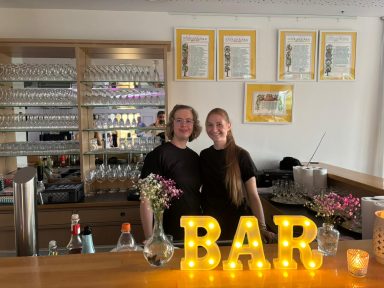 Zwei Personen stehen hinter einer Bar mit leuchtenden Buchstaben "BAR" und Blumen.