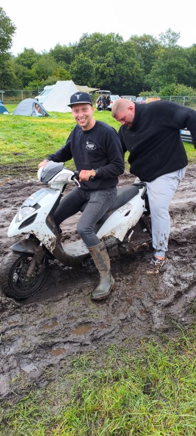 Zwei Männer auf einem weißen Moped in einem schlammigen Gelände, einer hilft dem anderen.