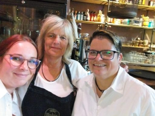 Drei Frauen lächeln in einem Café, eine trägt ein schwarzes Schürzenoutfit.