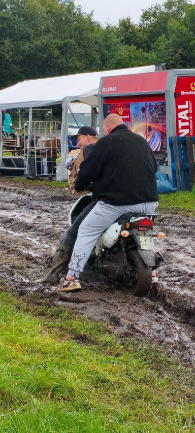 Zwei Personen sitzen auf einem Motorrad, das in schlammigem Gelände steht.