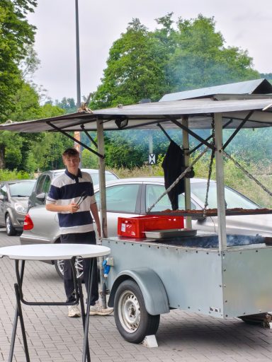 Ein Verkaufsstand mit einem Mann in gestreiftem Shirt, neben Autos und rauchender Grillfläche.