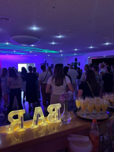 Party mit vielen Menschen, leuchtenden "BAR"-Buchstaben und farbigem Licht im Hintergrund.