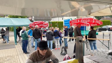 Menschen stehen in einer Reihe vor Ständen, mit roten Sonnenschirmen im Hintergrund.