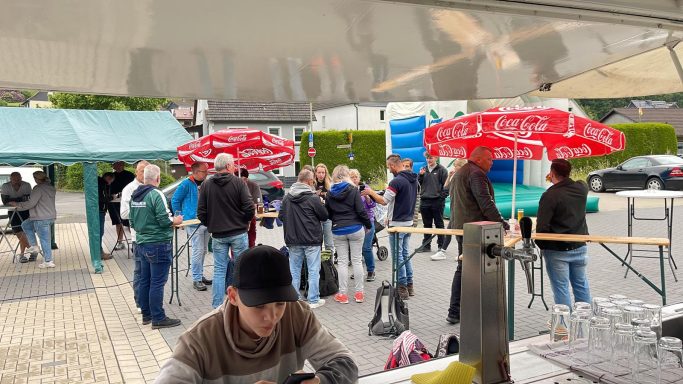Menschen stehen in einer Reihe vor Ständen, mit roten Sonnenschirmen im Hintergrund.