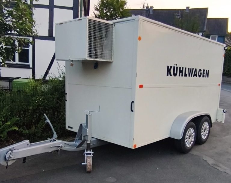 Weißer Anhänger mit zwei Achsen, beschriftet mit "KÜHLWAGEN". Steht auf einer Straße.