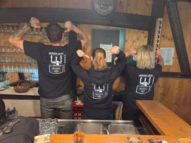 Drei Personen in schwarzen T-Shirts zeigen mit den Händen nach oben, Rücken zur Kamera.