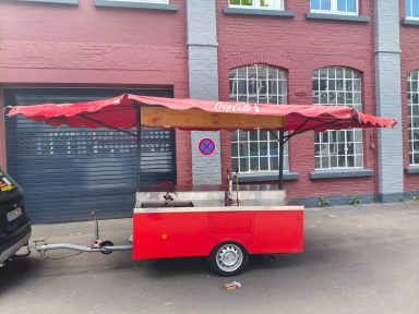 Roter Verkaufswagen mit rotem Sonnenschutzdach vor einem Gebäude.